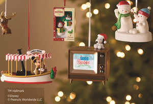 A holiday ornament display featuring multiple Peanuts-themed Christmas ornaments hanging against a warm, blurred Christmas tree background with bokeh lights. The ornaments include a Mickey Mouse carousel ride scene, a retro television with Snoopy and Woodstock, a snowman on a base labeled 'Peanuts Christmas', a packaged ornament, a reindeer, and a small hanging ornament.