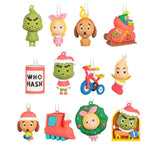 A set of colorful Christmas-themed character ornaments arranged on a plain white background. The ornaments include a green Grinch-like figure, a pink-haired girl in pink, a brown dog with a gold hat, a red Santa sack, a red can labeled 'WHO HASH', a green Grinch figure in a red Santa outfit, a red tricycle, a pink baby figure in a red stocking, a red train, a green wreath with a pink-haired girl, and a yellow Grinch figure in a window scene.